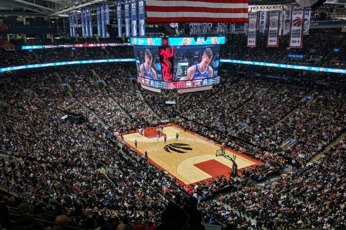 A crowded basketball arena with a large American flag hanging above the court